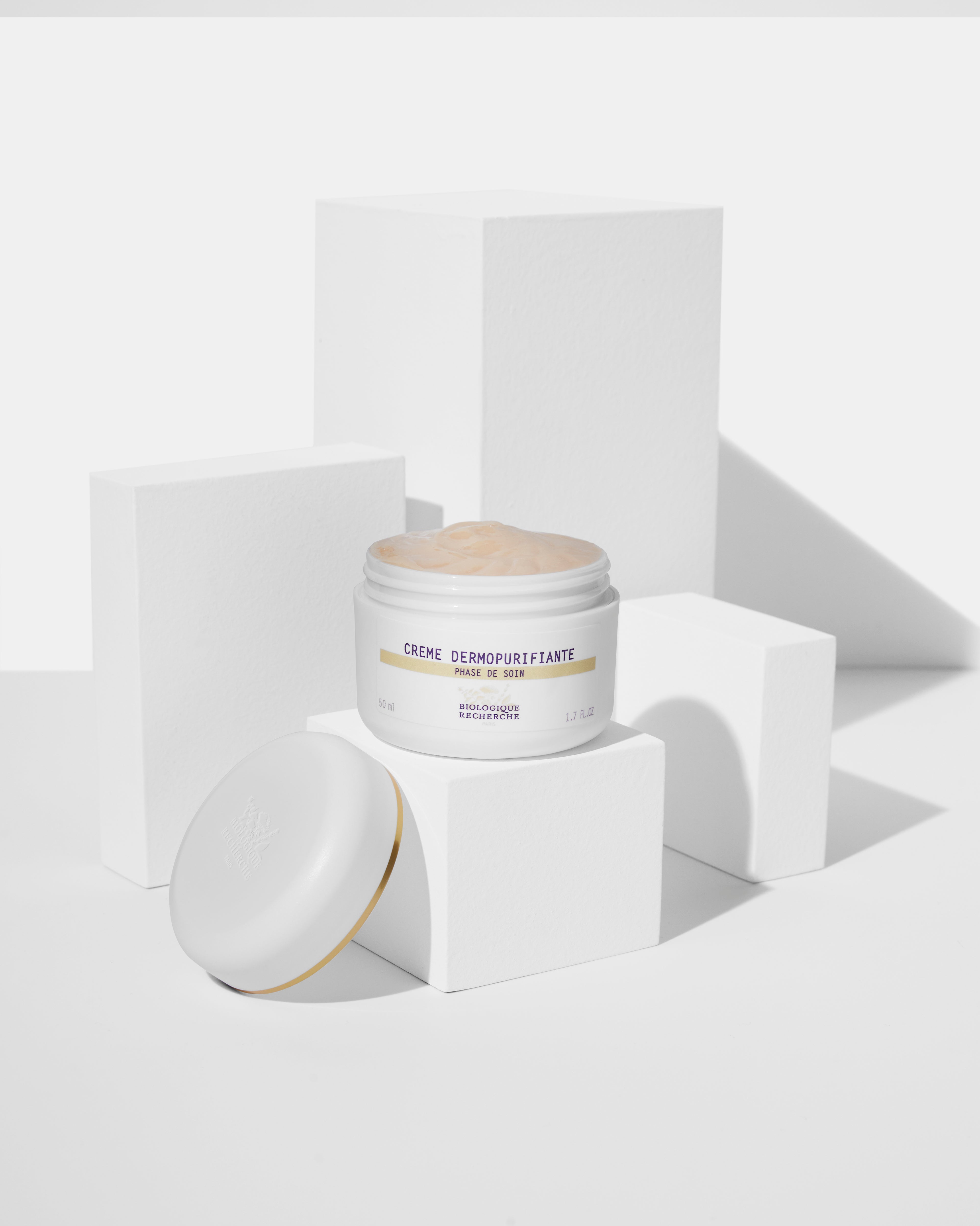 Open white jar of Biologique Recherche Crème Dermopurifiante cream with white lid beside it on white geometric blocks.