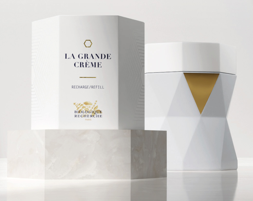 White hexagonal jar with gold triangular accent beside matching box labeled La Grande Crème on marble pedestal.
