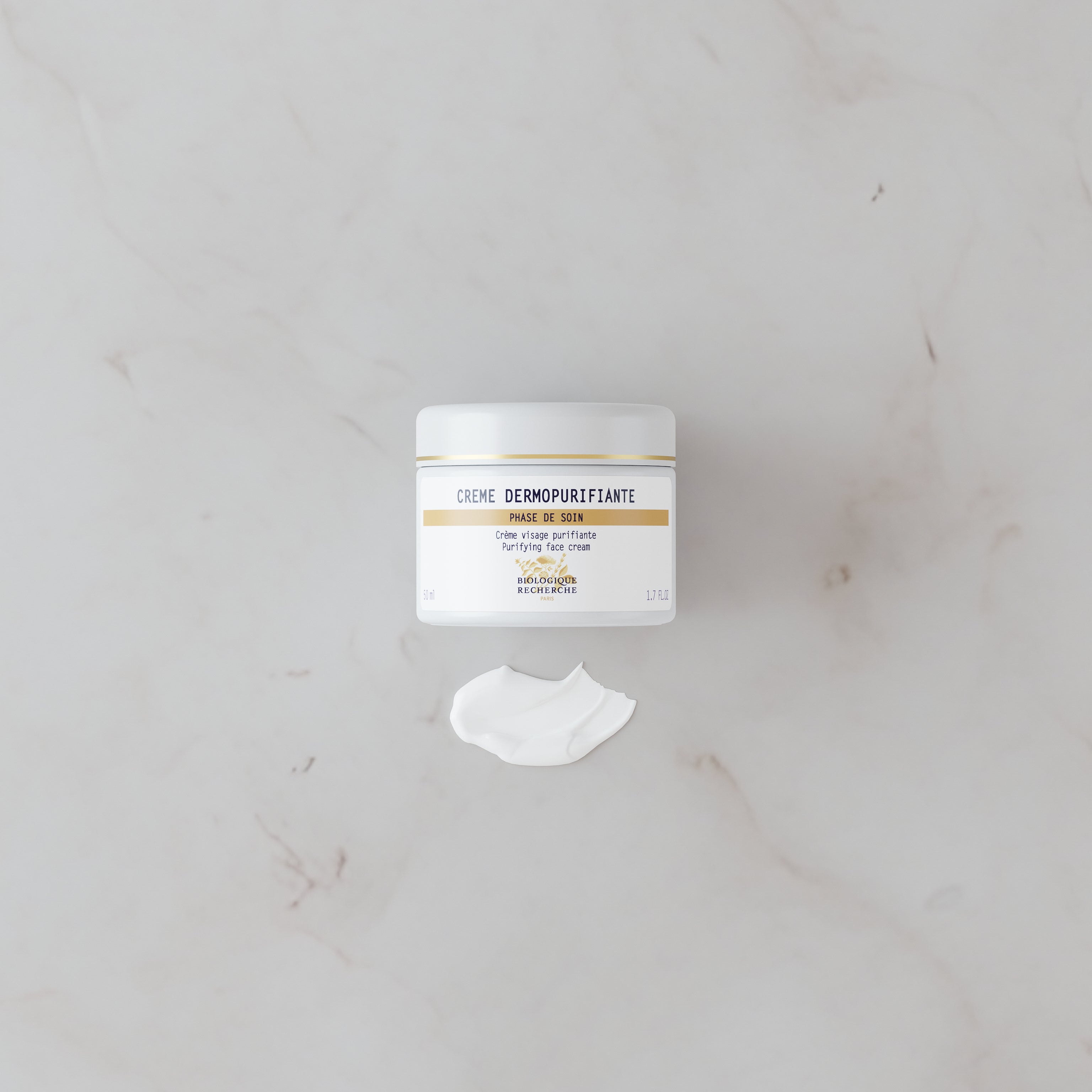 White jar of Crème Dermopurifiante with a dollop of cream on a light marble surface.