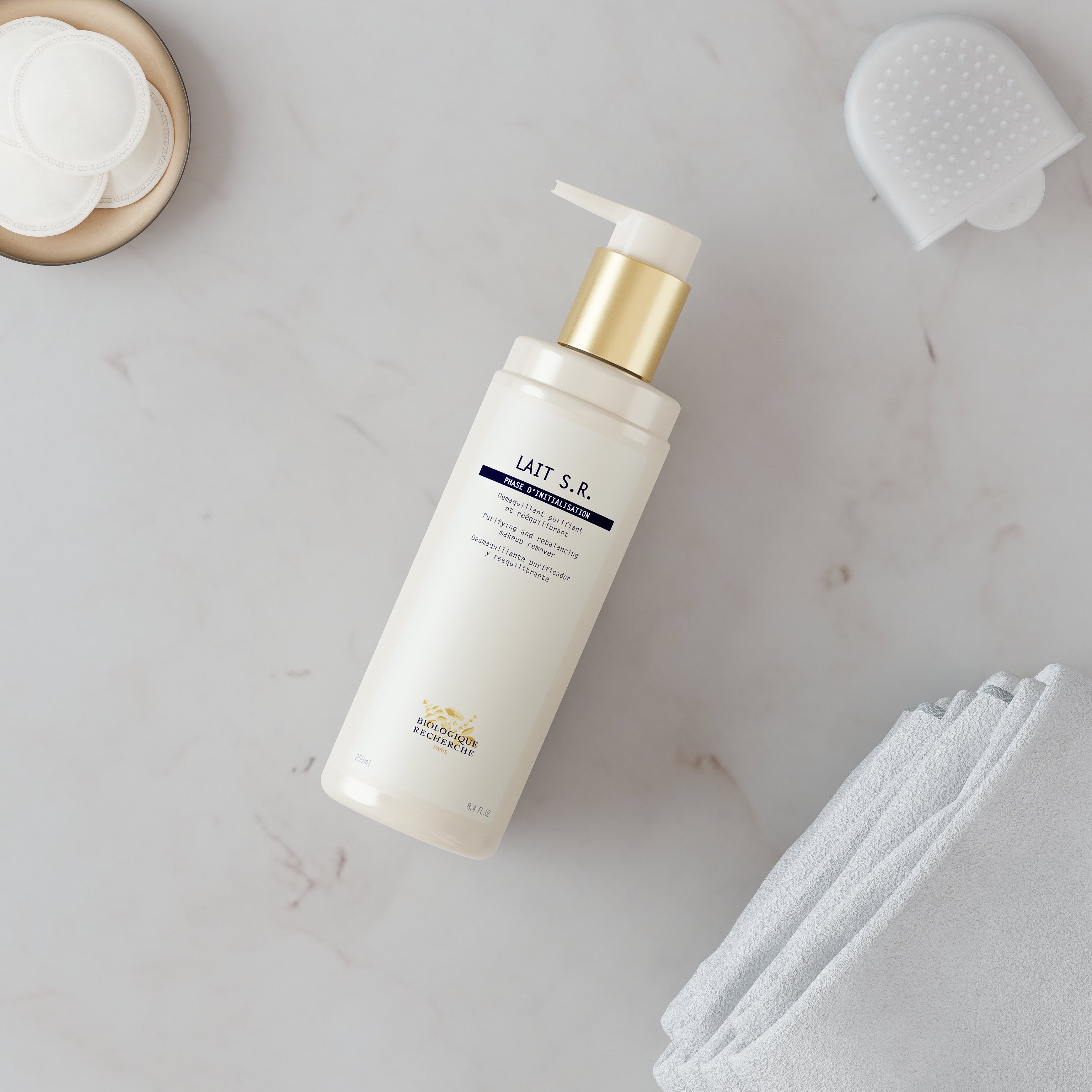 White plastic pump bottle of Lait S.R. cleanser with gold pump on marble surface next to white towel and skincare accessories.