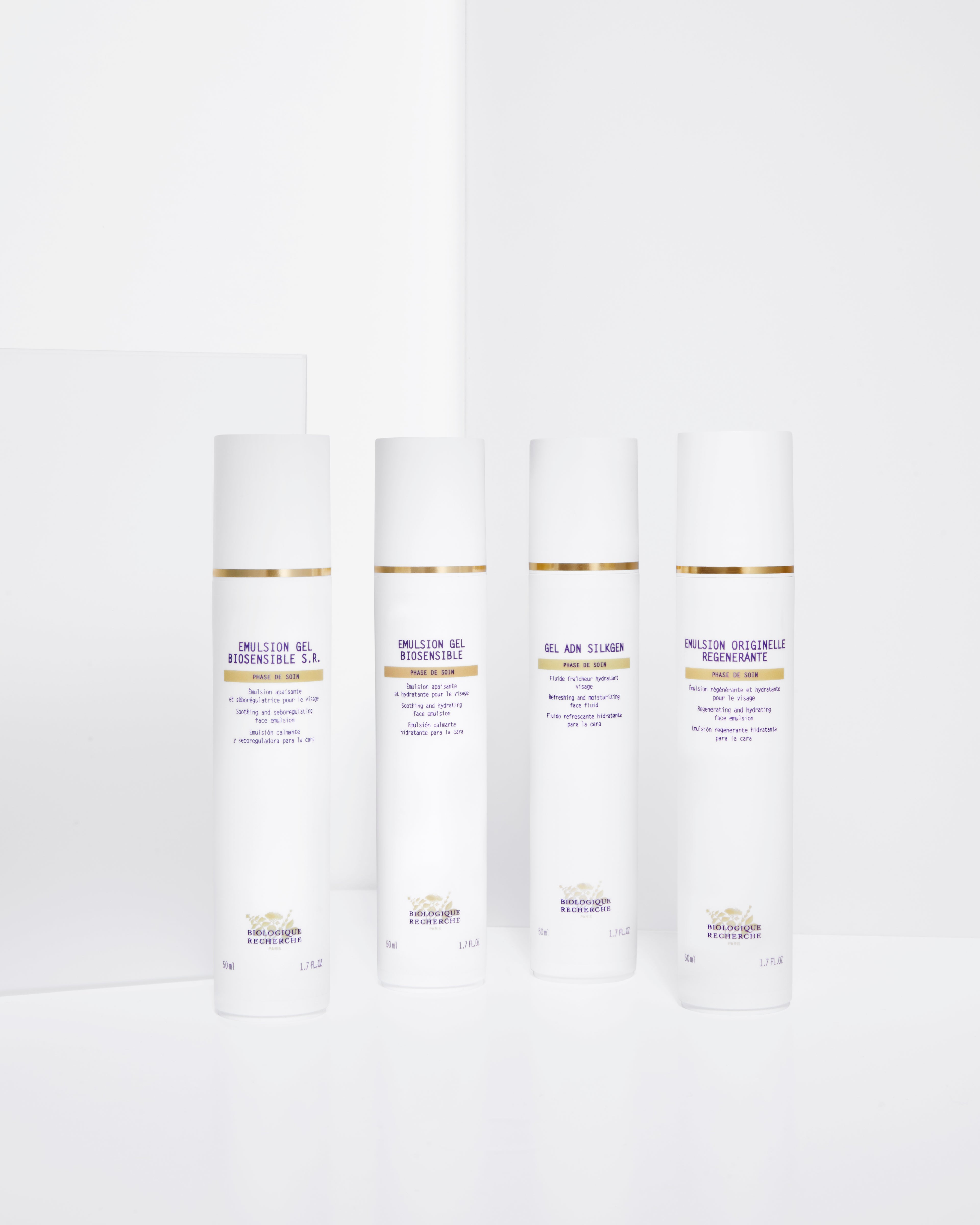 Four tall white cylindrical bottles of Emulsion Originelle Régénérante with gold accents and purple text on a plain white.