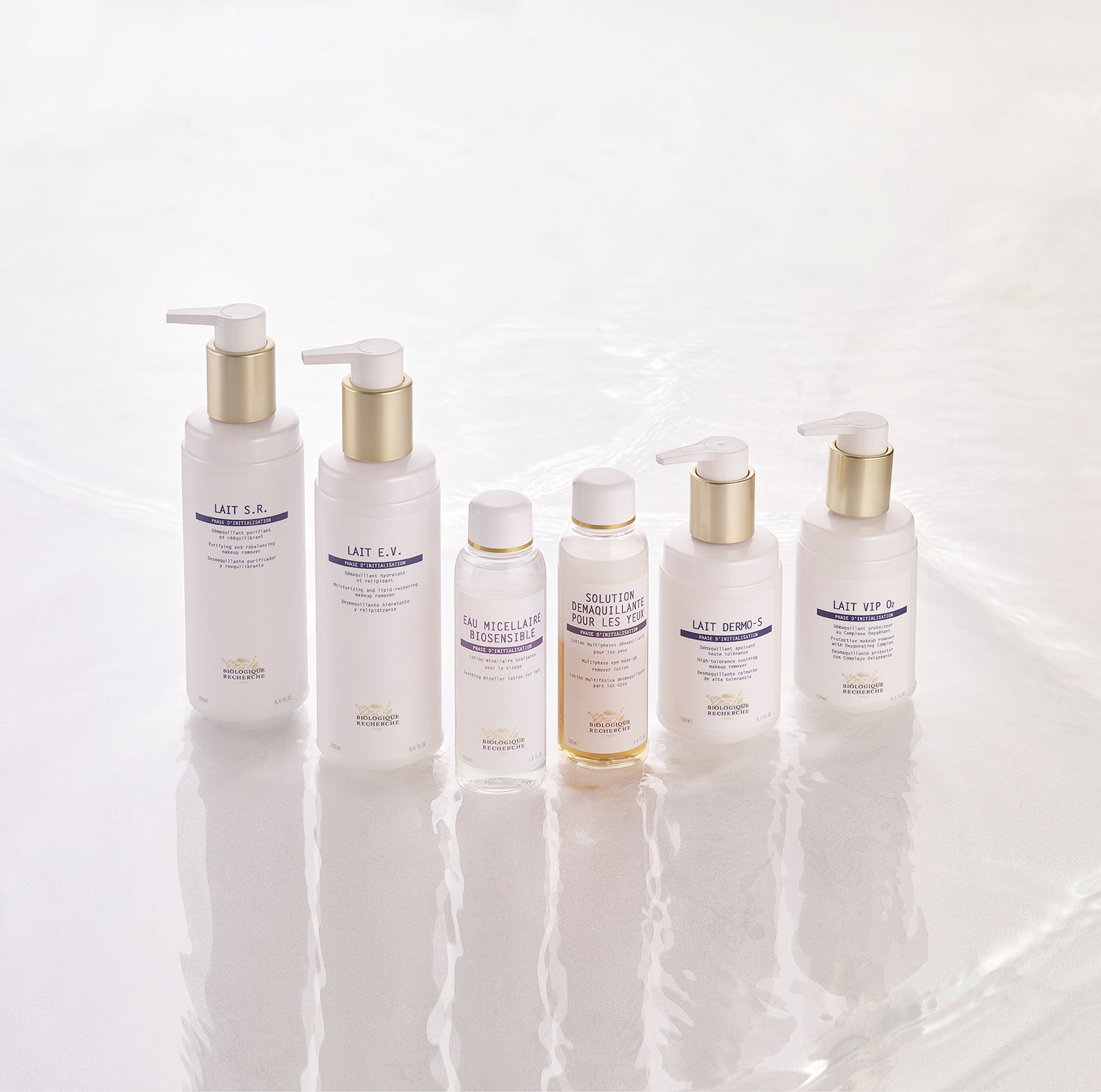 Six white cylindrical skincare bottles with gold and white caps, varying in size, arranged on a reflective surface.