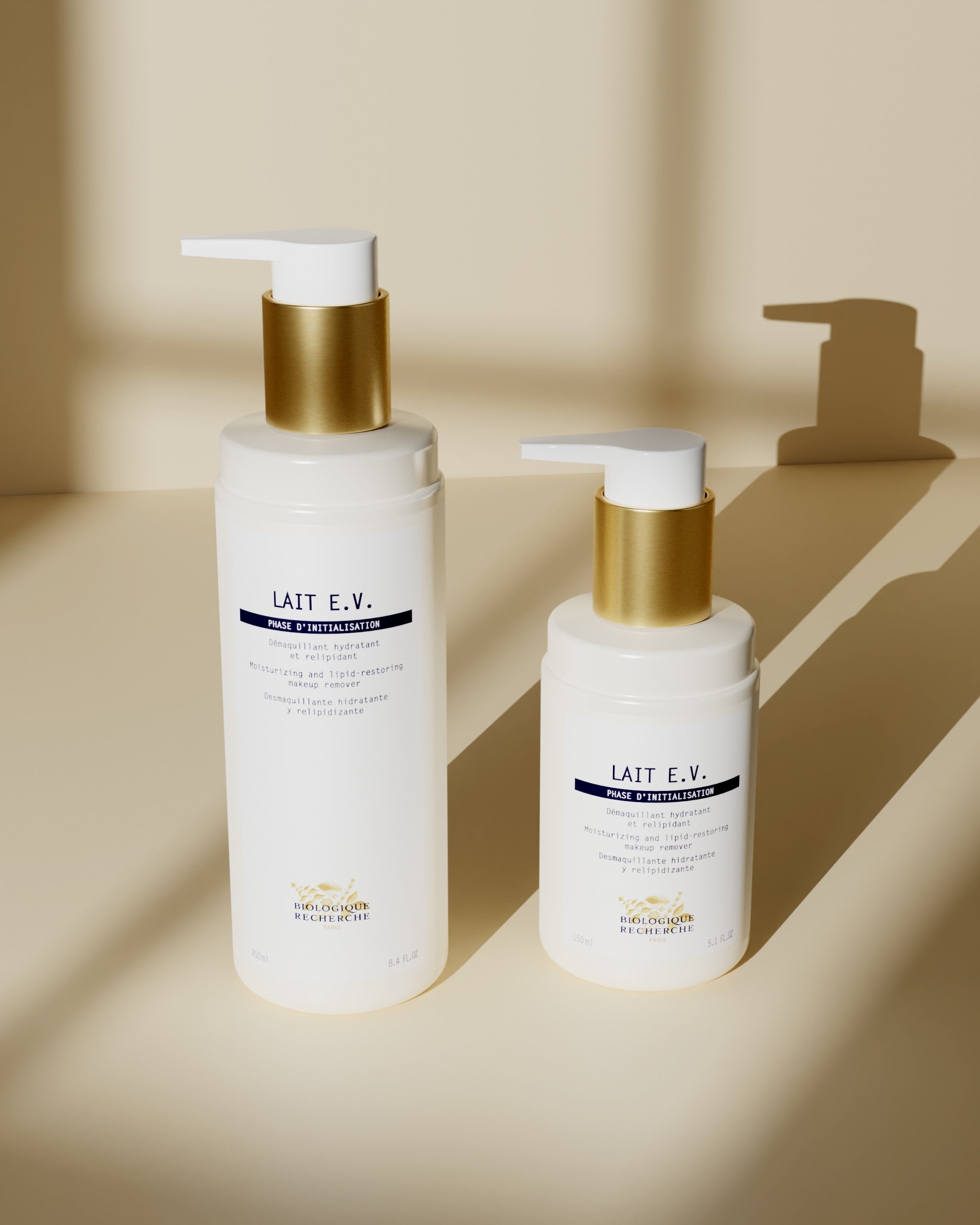 Two white plastic bottles of Lait E.V. milk cleanser with gold pump caps, one taller 250ml and one shorter 150ml, on beige.