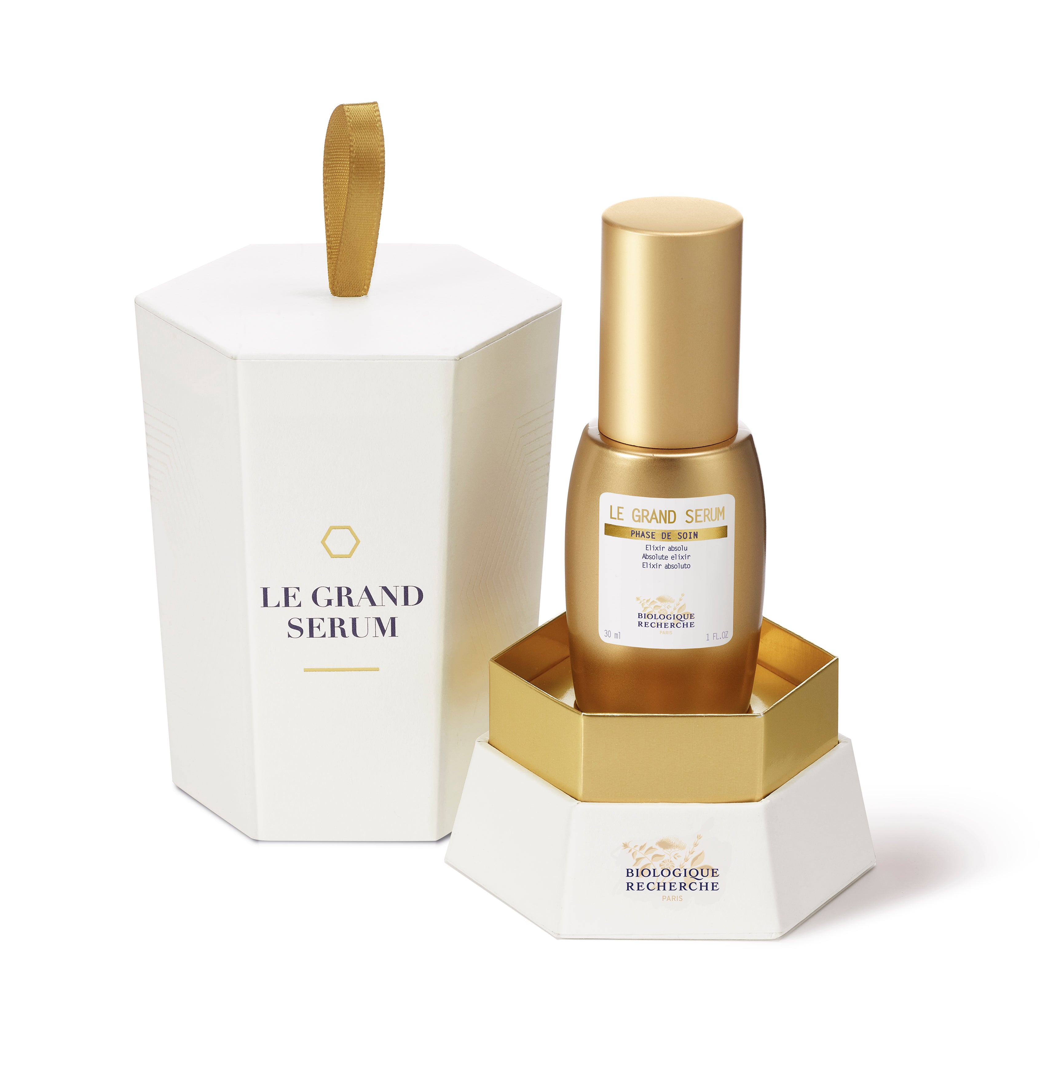 Le Grand Sérum bottle with gold cap and hexagonal white and gold packaging box with ribbon handle.