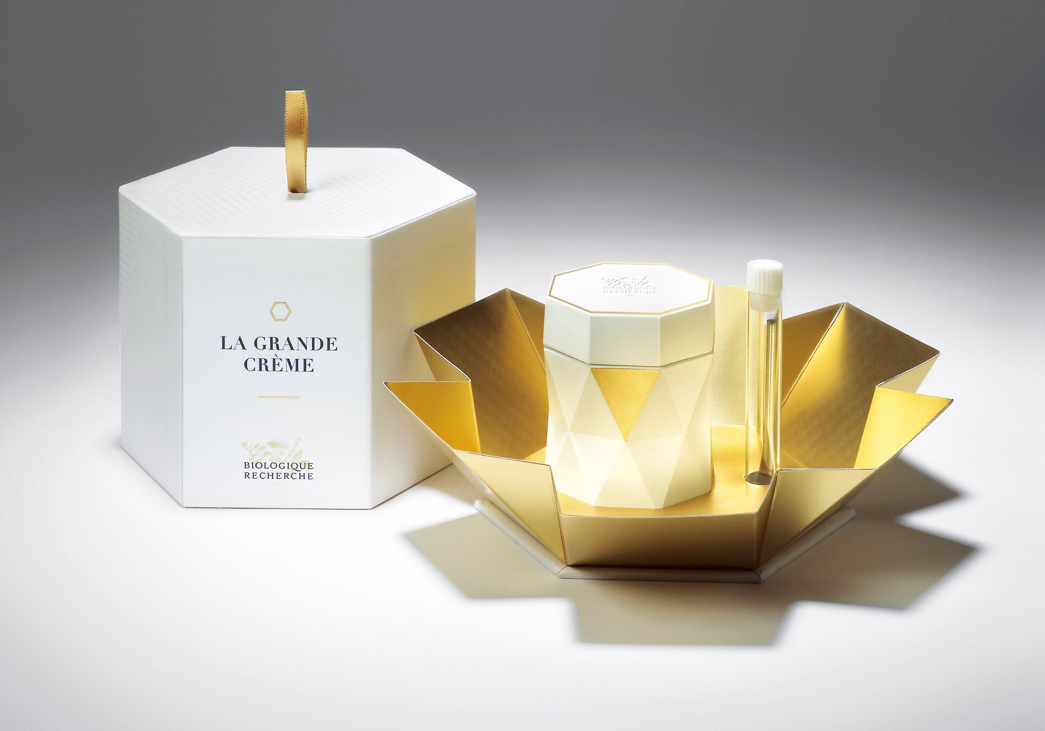 Hexagonal white and gold jar of La Grande Crème with white and gold geometric box and open gold folding case.