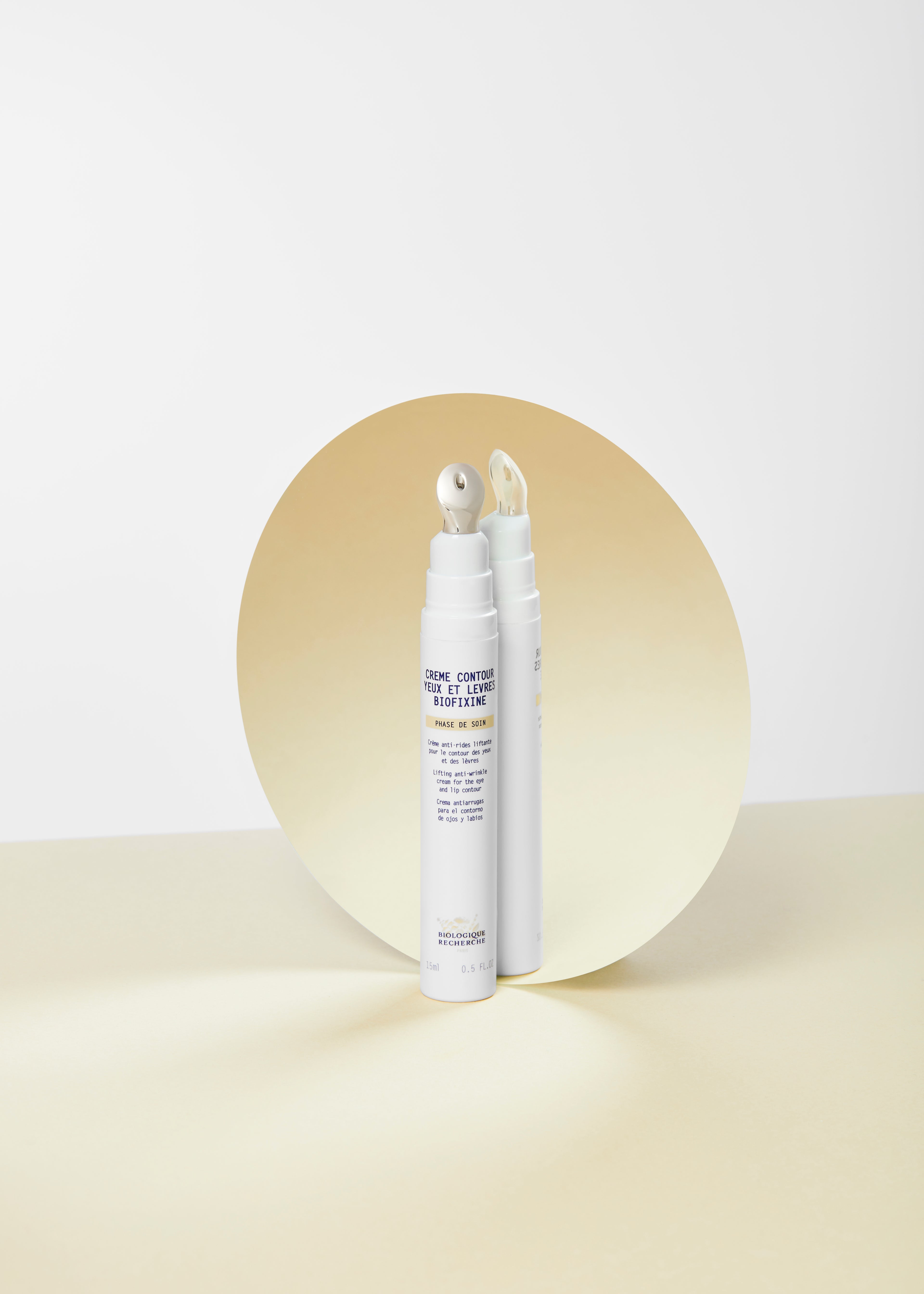 White slim tube of Crème Contour Yeux et Lèvres Biofixine with roller applicator on round golden reflective background.