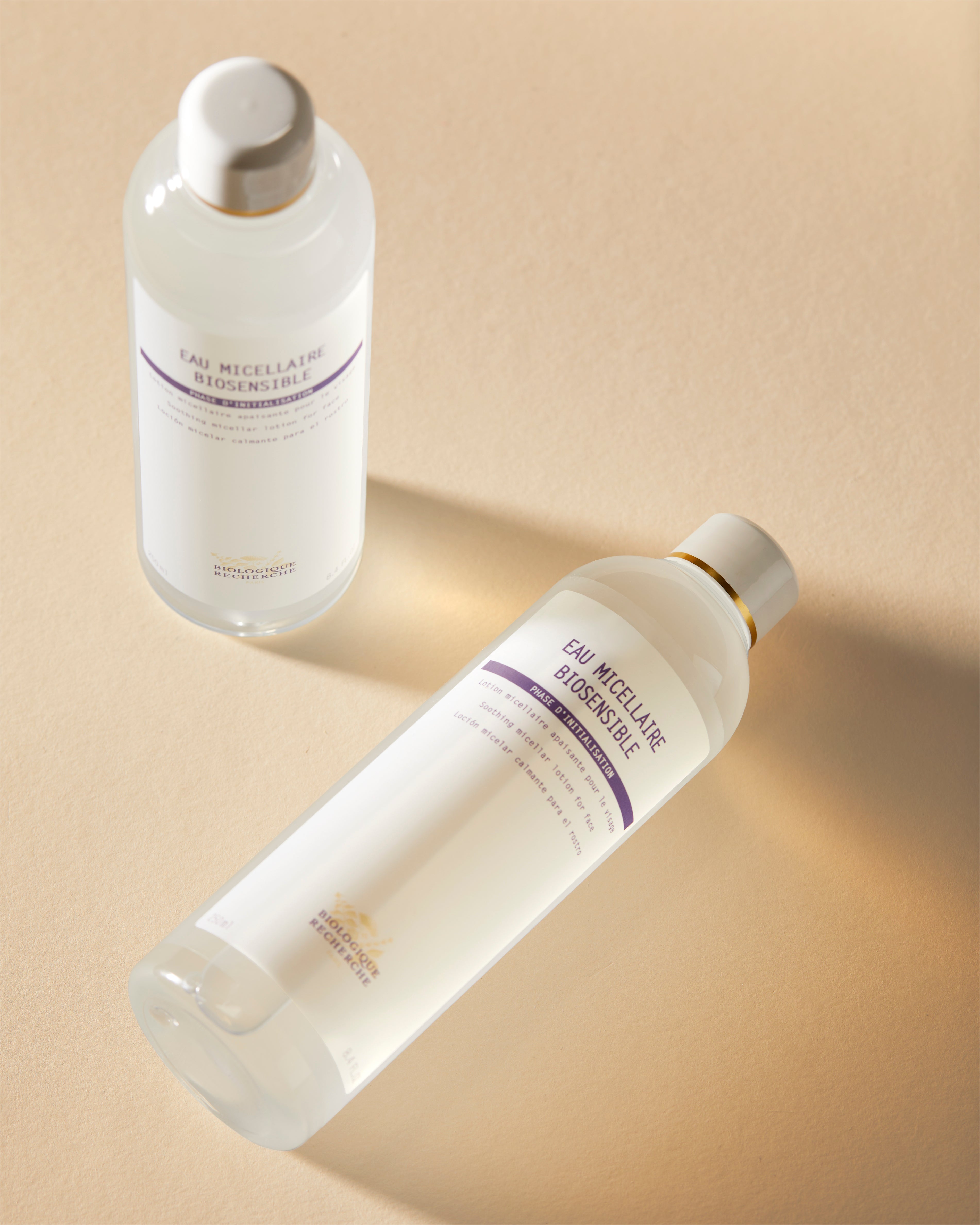 Two clear bottles of Eau Micellaire Biosensible cleanser with white labels and beige caps on a beige surface, one standing.