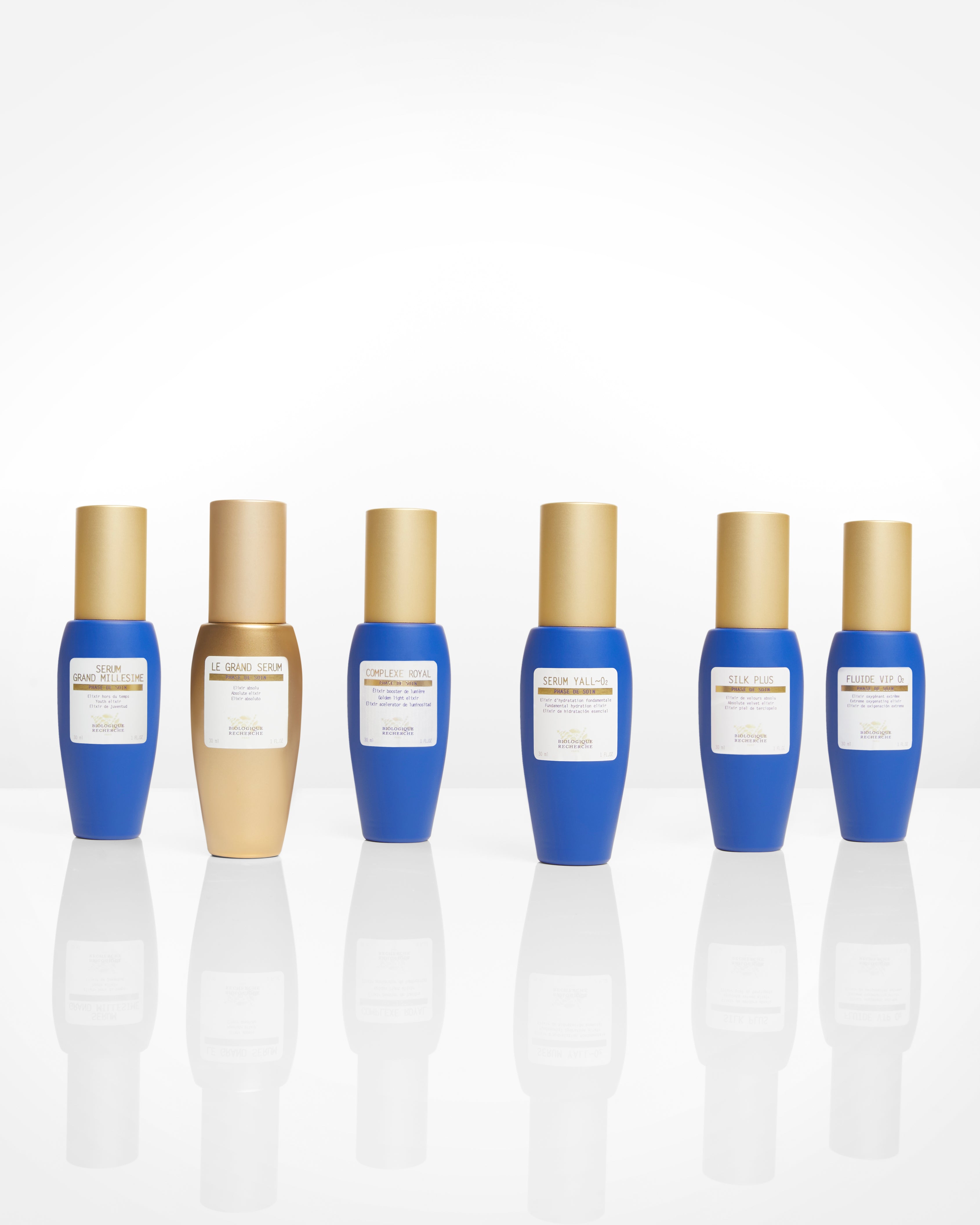 Six Le Grand Sérum bottles in blue and beige with gold caps, labeled for skincare use.