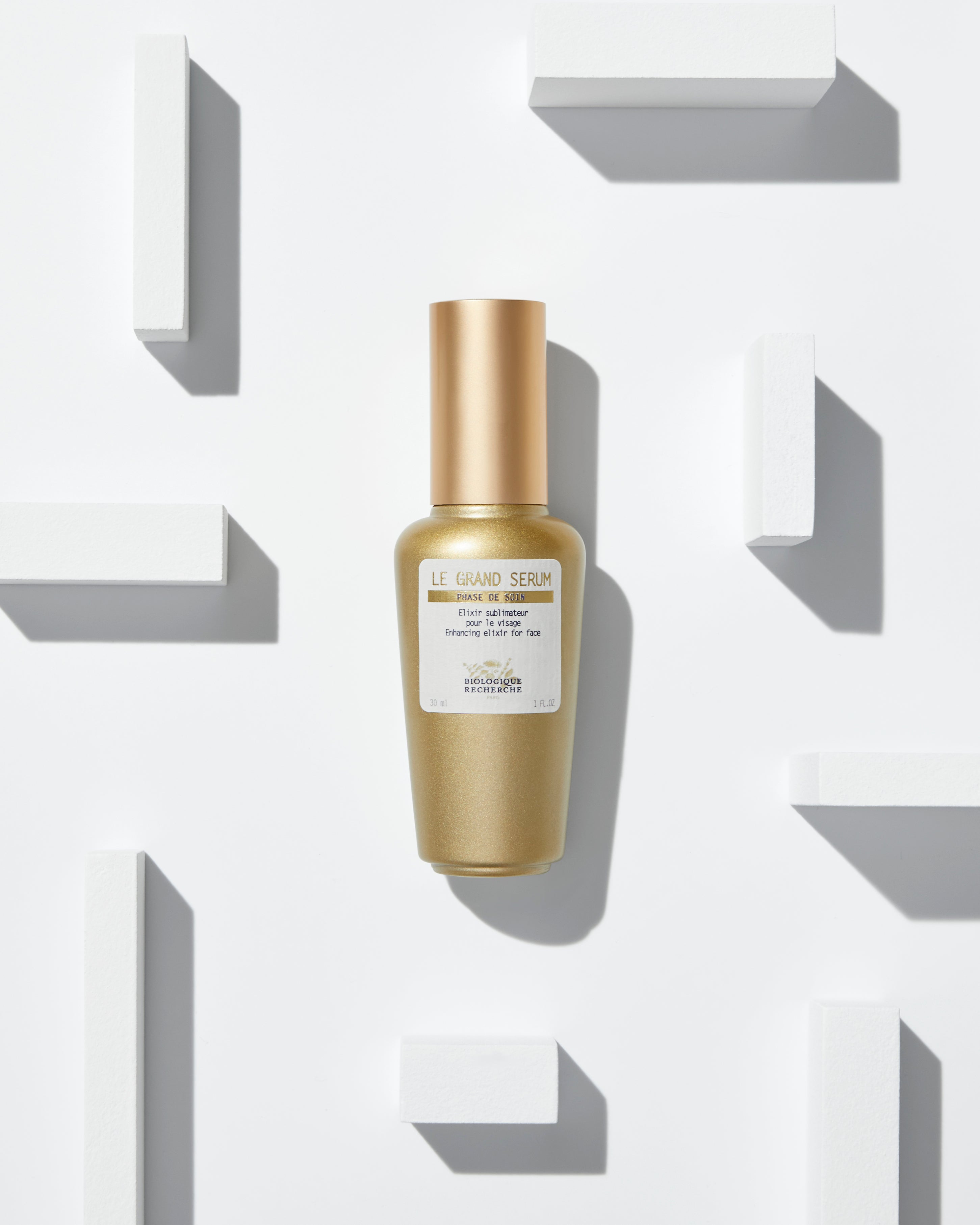 Gold metallic bottle of Le Grand Sérum with white label and gold cap on white geometric background.