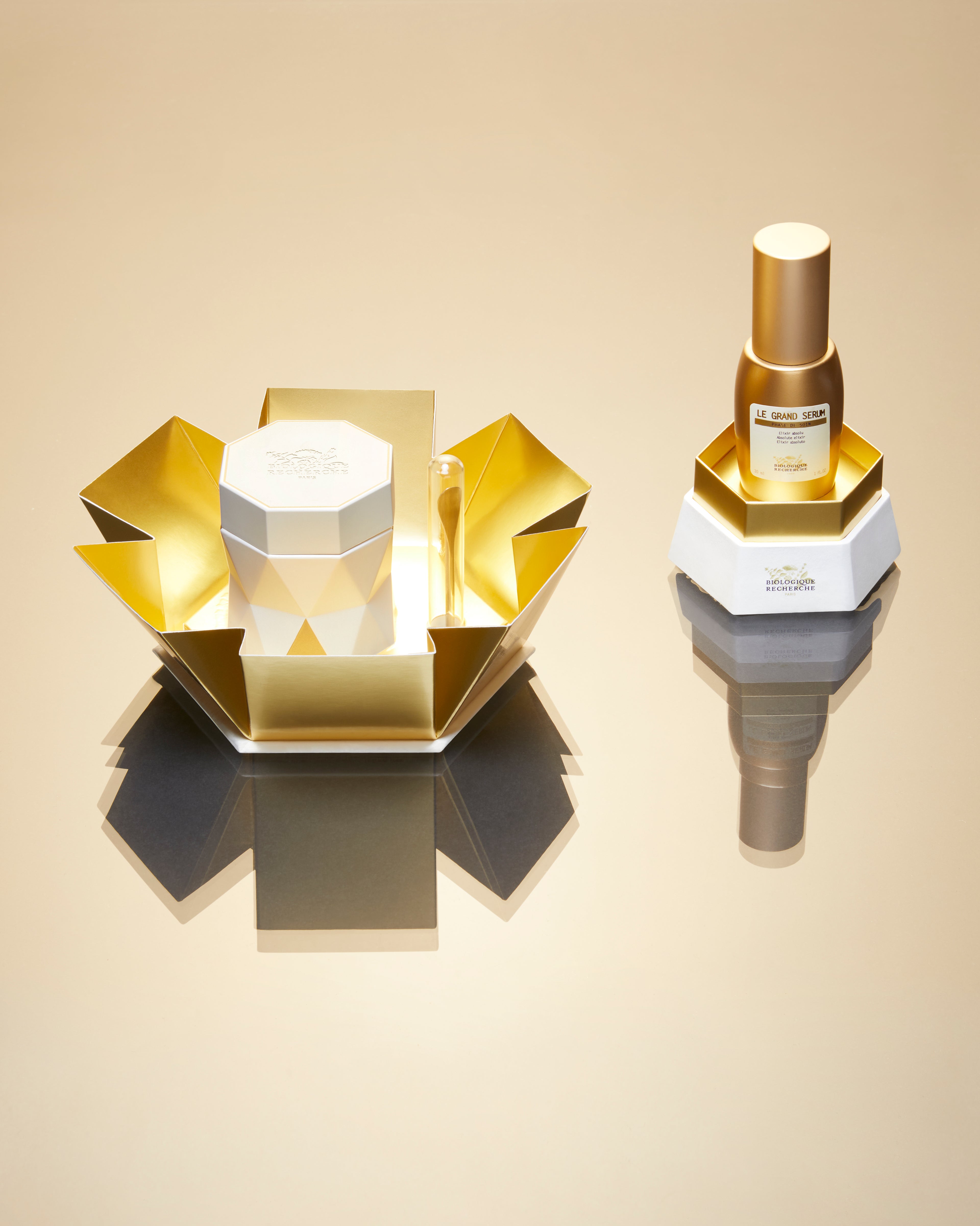 Hexagonal white jar with gold lid beside matching gold and white box on reflective surface.