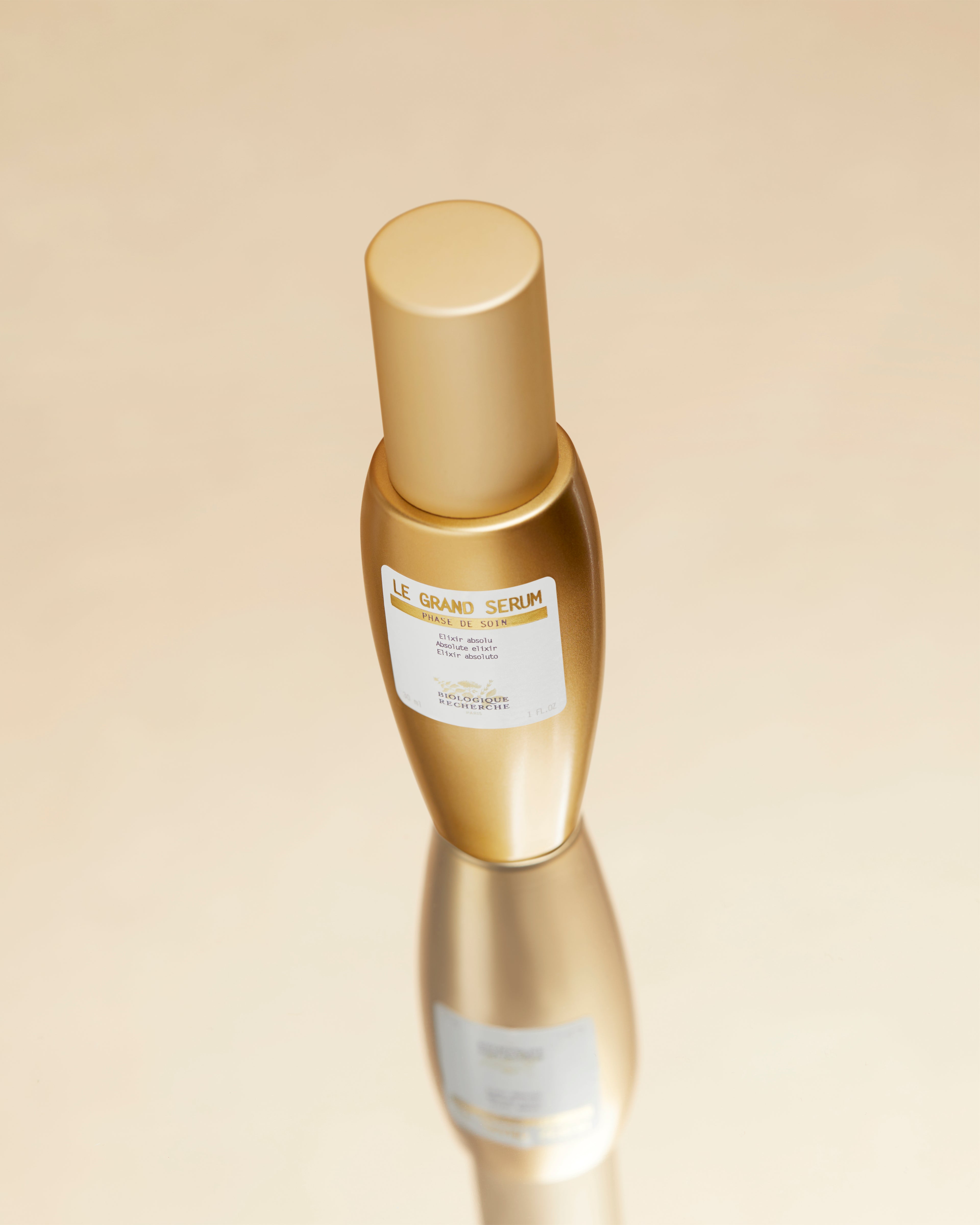 Gold cylindrical bottle with a beige label and cap reflecting on a light beige surface.