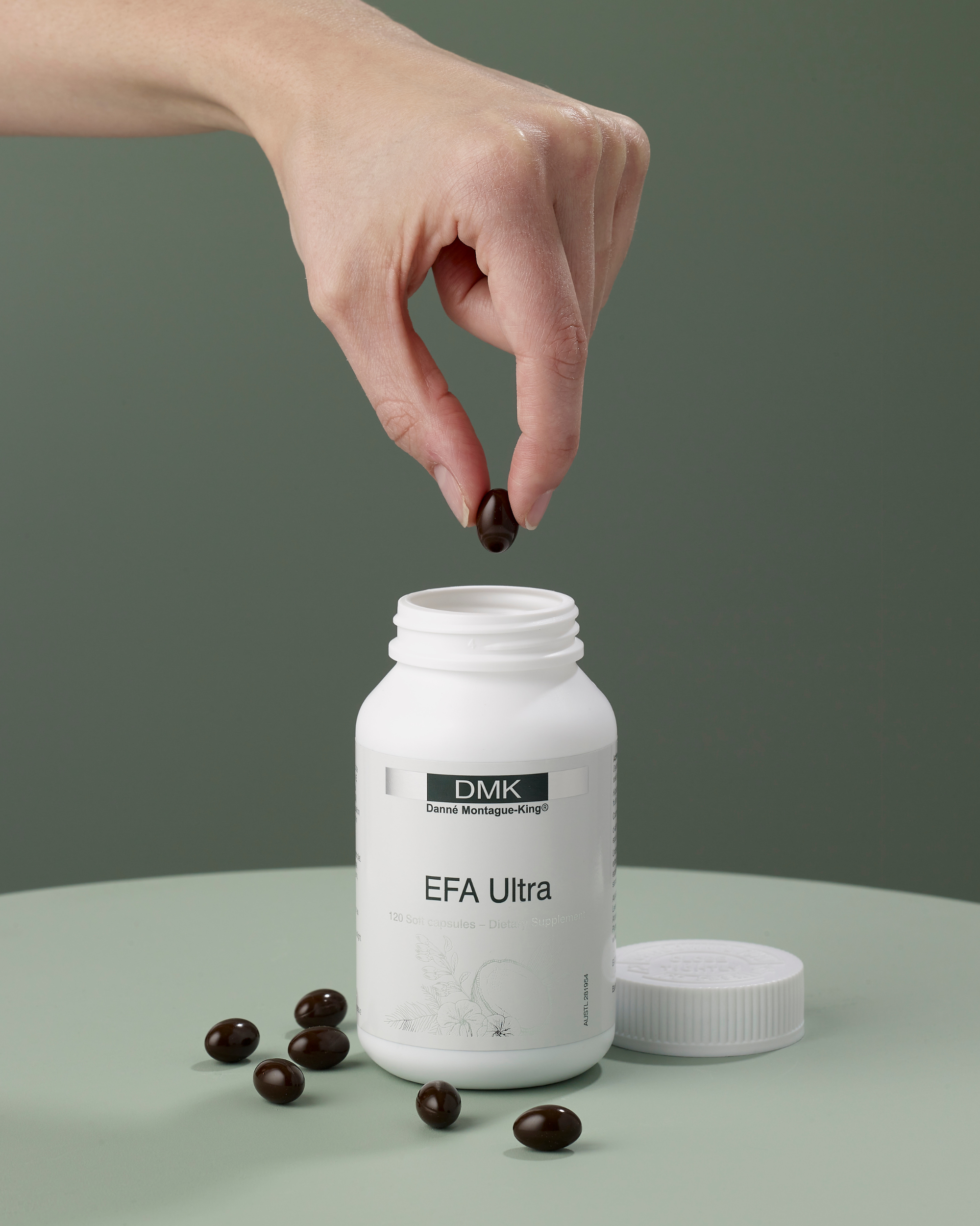 White supplement bottle labeled EFA Ultra with black capsules, hand dropping one capsule in focus on green background.