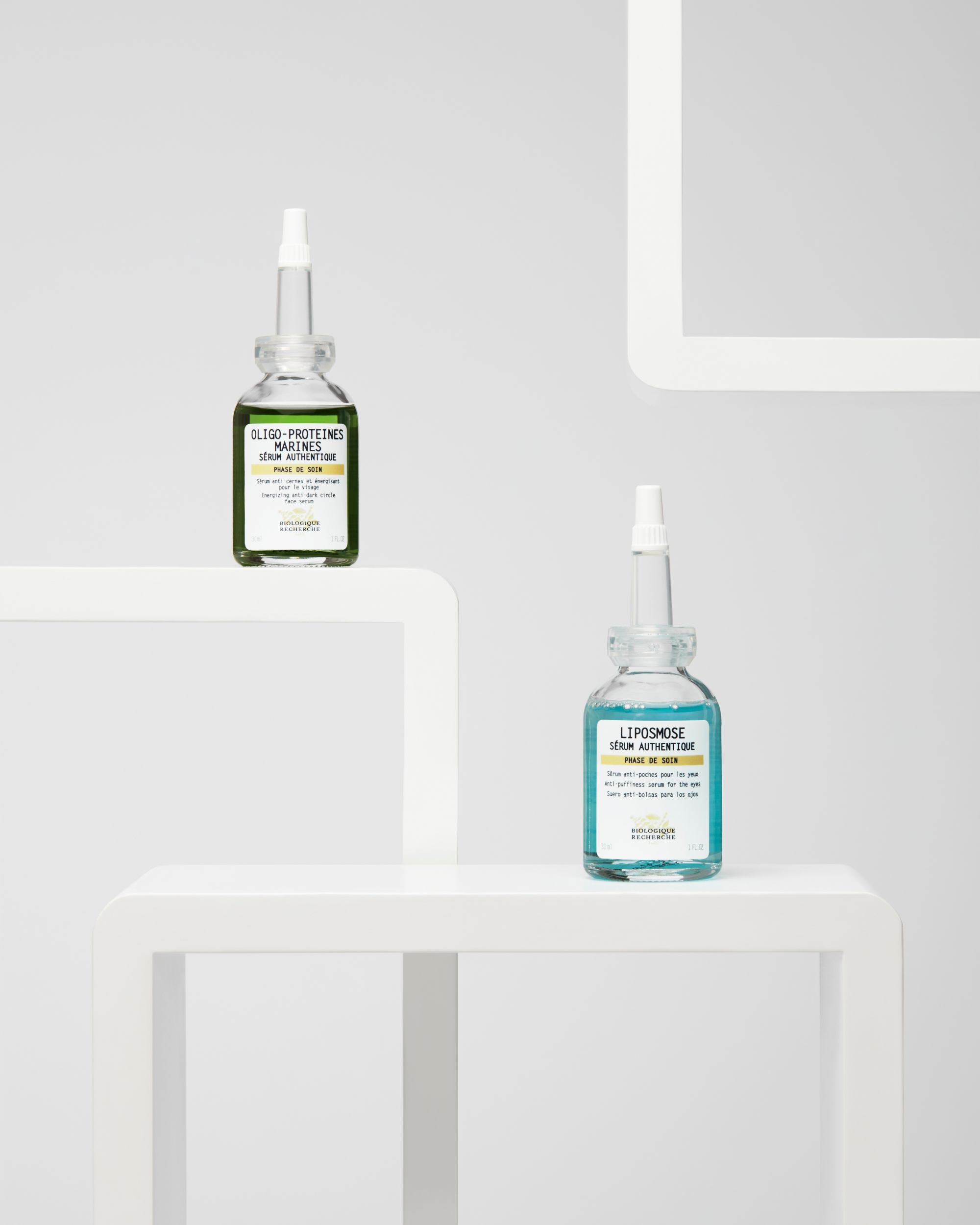 Two small glass bottles of Biologique Recherche Liposmose eye care serum, one with green liquid and one with blue liquid,.