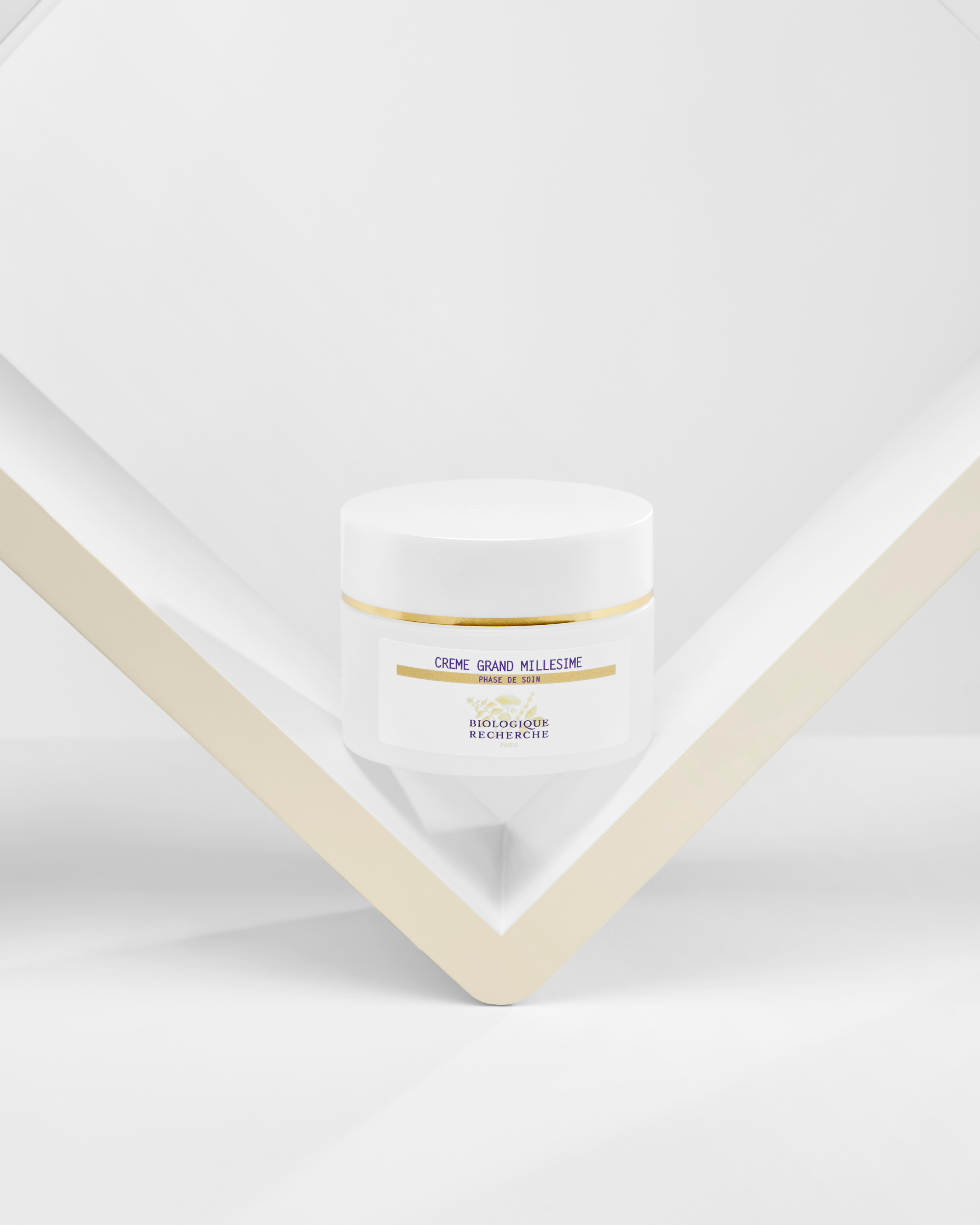 White jar of Crème Grand Millésime with gold and purple label, placed on an angled beige frame against a white background.