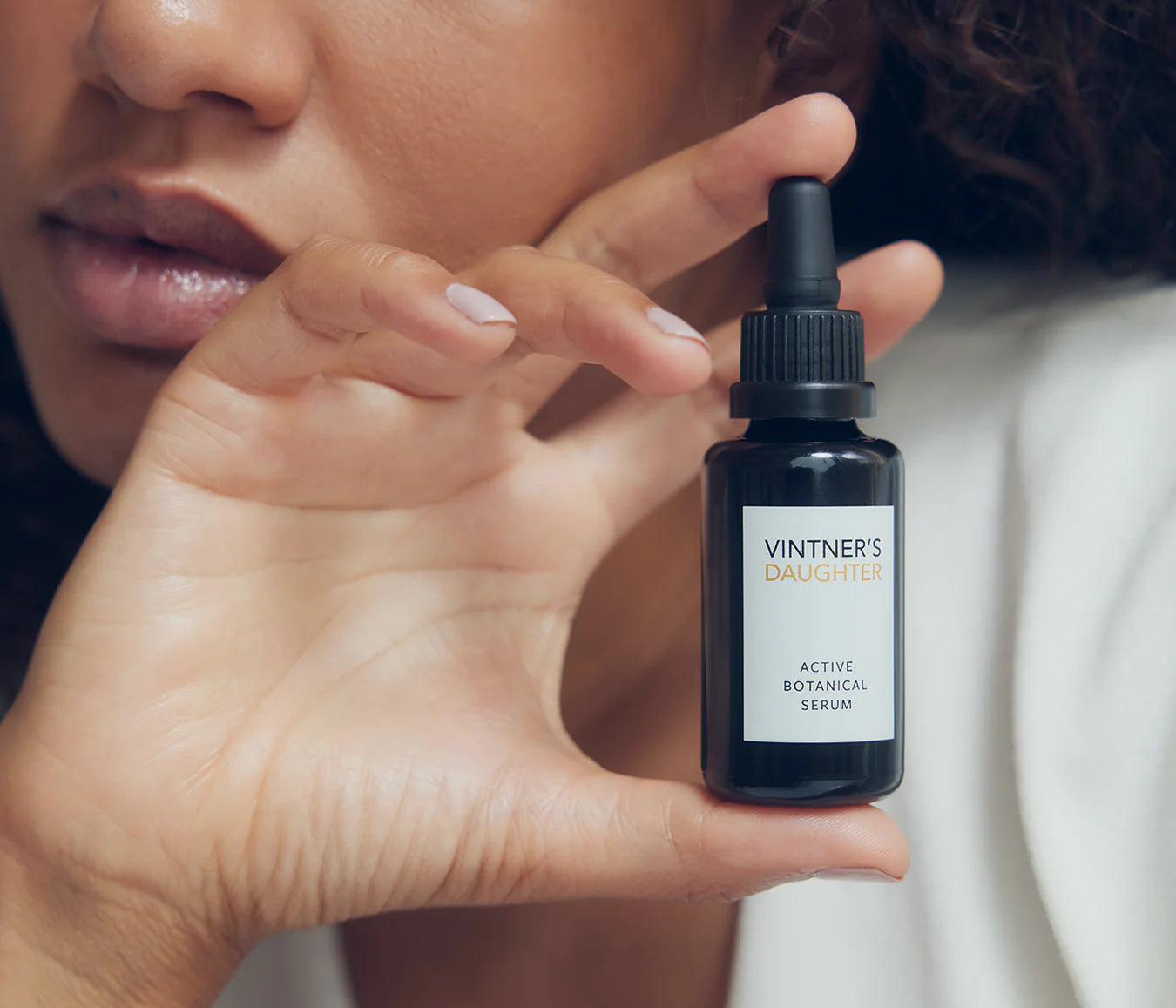 Dark amber glass dropper bottle labeled Active Botanical Serum held by a person with a light brown complexion and curly hair.